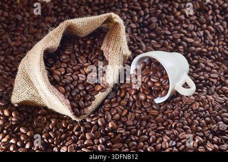 Grains de café torréfiés se renversant d'un sac en toile de jute et d'une tasse blanche, créant une texture de fond marron riche Banque D'Images