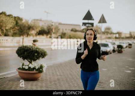 Une femme fait du jogging dans une rue à Malte. Elle porte des écouteurs et court devant des magasins et des voitures garées. Le soleil brille et le ciel est clair, indicat Banque D'Images