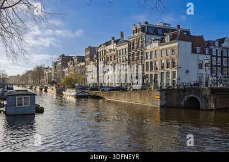 Vue pittoresque sur le canal Herengracht à Amsterdam avec des maisons marchandes historiques, une péniche et un pont de pierre sous un ciel bleu. Banque D'Images