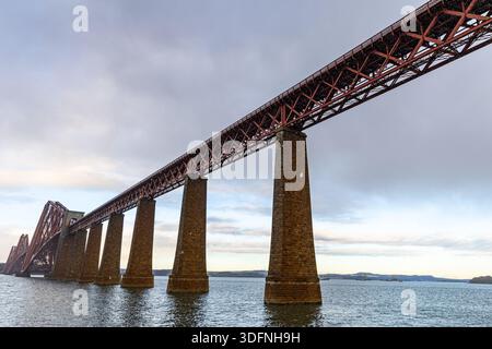 Un plan large et à angle bas d'un grand pont ferroviaire rouge avec des supports en pierre s'étendant au-dessus de l'eau sous un ciel nuageux. Banque D'Images
