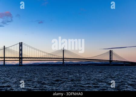 Un large pont suspendu s'étend sur une étendue d'eau sombre et agitée sous un ciel dégradé de bleus et d'oranges douces, avec des collines lointaines silhouettes. Banque D'Images