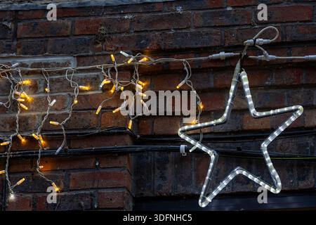 Un contour d'étoile de fil éclairé avec des LED blanches est suspendu contre un mur de briques, orné d'une chaîne de lumières LED jaunes chaudes. Banque D'Images