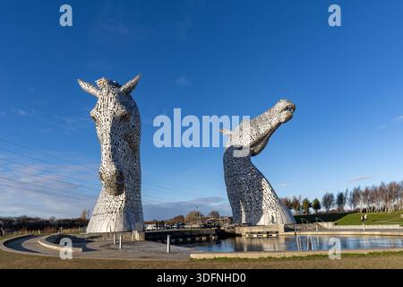 Deux imposantes sculptures de tête de cheval en acier inoxydable se dressent près d'un canal sous un ciel bleu vif, avec un paysage herbeux et des arbres en arrière-plan. Banque D'Images