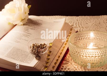 Bible ouverte montrant l'écriture avec un chapelet, une fleur blanche, et allumée bougie sur une nappe de dentelle Banque D'Images