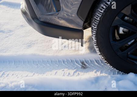 Pneu du véhicule et coin pare-chocs en conditions hivernales, roue noire avec bande de roulement enneigée et traces de piste fraîches sur la surface enneigée glacée à l'extérieur en plein jour Banque D'Images