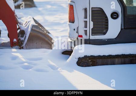 Mini-chargeuse à chenilles recouverte de neige stationnée dans la neige profonde, vue rapprochée de la chenille en caoutchouc et du panneau de carrosserie arrière avec godet de pelle hydraulique flou en hiver Banque D'Images