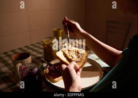 Une personne étale du beurre d'arachide sur du pain grillé pour un petit déjeuner au beurre d'arachide et à la gelée. Confiture, tasse et une table à carreaux créent une ambiance chaleureuse dans la cuisine. Banque D'Images