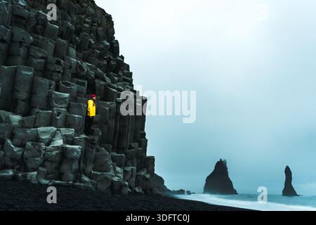 Vue d'une figure solitaire dans une veste jaune vif perchée sur des colonnes de basalte, contrastant avec le sable sombre et les piles de mer brumeuses, Vik, Islande. Banque D'Images
