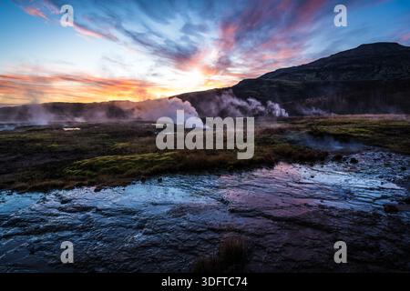 Vue de l'activité géothermique qui vaporise depuis le sol, reflétant le coucher de soleil coloré sur le paysage dans la vallée de Haukadalur, Haukadalur, Islande. Banque D'Images