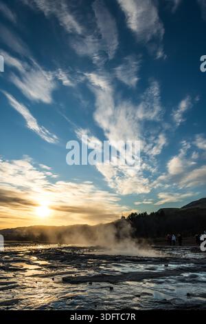Vue du spectacle géothermique avec un horizon enflammé dans les teintes flamboyantes du coucher du soleil, vapeur qui monte sur une toile de bleu et de nuages, vallée de Haukadalur, Islande. Banque D'Images