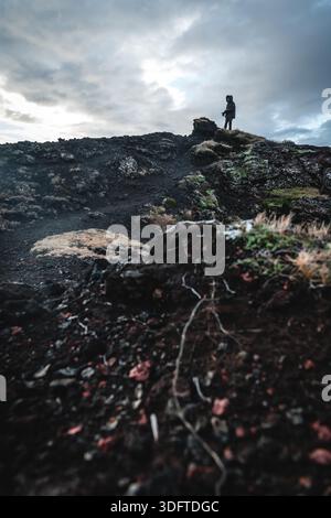 Vue d'une figure solitaire debout au sommet d'une formation rocheuse volcanique accidentée et sombre sous un ciel spectaculaire près du parc géothermique de Keflavik, Islande. Banque D'Images