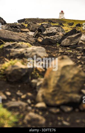 Vue d'un phare avec un sommet rouge perché au sommet d'une colline, surplombant un paysage rocheux avec des taches de vert, sous un ciel silencieux, Vik, Île du Sud, IC Banque D'Images