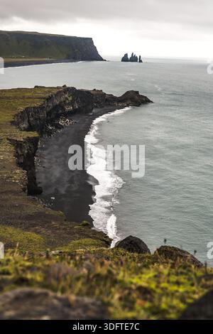 Vue sur la plage de sable noir embrassée par des vagues blanches mousseuses contrastant avec les falaises escarpées sous un ciel sombre, Vik, Île du Sud, Islande. Banque D'Images