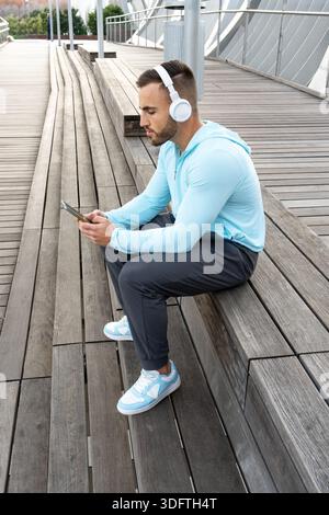 Un jeune homme se détend sur des bancs en bois sur une terrasse extérieure, écoutant de la musique à travers des écouteurs tout en étant concentré sur son smartphone. Le soleil brille bri Banque D'Images