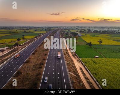 Vue aérienne de véhicules se déplaçant sur l'autoroute coupant à travers un paysage vibrant de champs de moutarde sous un ciel doré, Alwar, Rajasthan, Inde. Banque D'Images