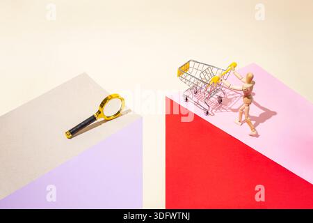 Photographie de style pop art d'un mannequin en bois poussant un chariot et une loupe sur fond de papier rose , rouge et beige disposés comme un Banque D'Images