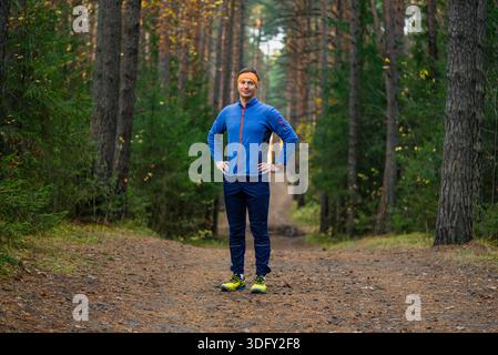 Homme en veste bleue et pantalon sombre debout avec les mains sur les hanches sur un sentier forestier sinueux, portant un bandeau orange et des baskets jaunes, incarnant le fitness, la santé et l'activité de plein air Banque D'Images