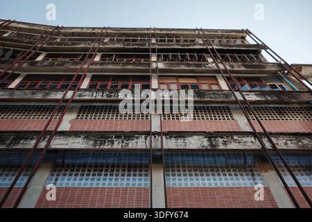 Une perspective à faible angle d'une façade de bâtiment vieillie de plusieurs étages avec poutres en béton altéré, sections de carrelage rouge, blocs de verre et rangées de trous Banque D'Images