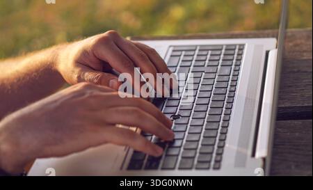 Gros plan des mains numériques nomades freelance tapant sur ordinateur portable. Gros plan de mains tapant sur un ordinateur portable à l'extérieur sur une table en bois. Banque D'Images