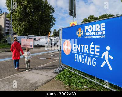 Strasbourg, France - SPE 14, 2025 : visiteurs mère et fils marchent vers l'entrée de la Foire européenne de Strasbourg devant une grande signalisation bleue guidant Pedest Banque D'Images