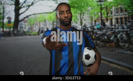 Jeune homme afro-américain en maillot bleu tient le ballon de football et montre un geste d'arrêt de paume dans la rue de la ville ; détermination de soutien de l'équipe. Banque D'Images