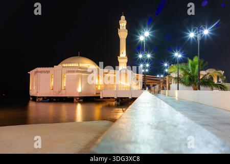 Djeddah, Arabie Saoudite - 23 décembre 2025 : une scène nocturne tranquille d'une mosquée blanche Al Rahma, mosquée flottante, sur pilotis au bord de l'eau, reliée à un L. Banque D'Images