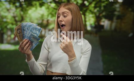 Jeune femme blonde dans un parc détenant des billets de banque hongrois en forint à l'extérieur, exprimant le bonheur et le succès financier, une verdure vibrante dans le backgrou Banque D'Images