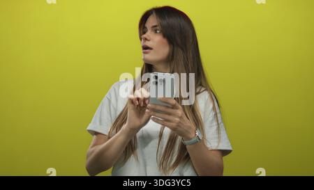 Jeune femme hispanique à l'aide d'un smartphone sur fond jaune vif portant un tshirt blanc décontracté, absorbé dans l'interaction technologique Banque D'Images