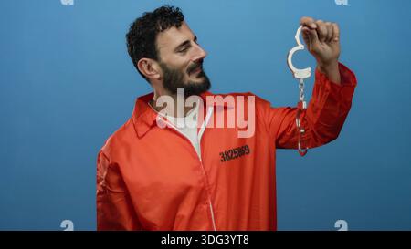 Homme hispanique avec la barbe dans la combinaison de prison orange tenant les menottes sur fond bleu, représentant le thème de l'incarcération. Banque D'Images