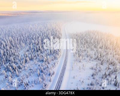 Vue aérienne d'un ruban de route qui traverse une forêt immaculée et enneigée sous une douce lumière éthérée, Laponie, Finlande. Banque D'Images