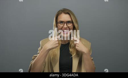 Femme souriant et faisant des gestes à elle-même dans un blazer beige sur un fond gris Uni. Banque D'Images