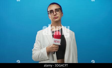 Femme journaliste tient le microphone d'une main, coulant et portant des lunettes et blazer blanc sur fond de studio bleu, mousse de micro rouge visible ; calme Banque D'Images