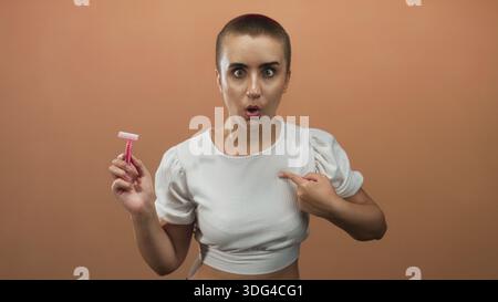 Femme aux cheveux courts tenant un rasoir jetable rose, pointe le doigt sur la poitrine tout en portant un haut blanc recadré en studio ; autonomisation. Banque D'Images