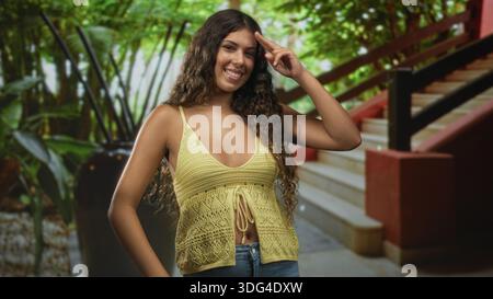 Femme sourit, montre un décolleté et lève deux doigts tout en portant un haut en tricot jaune et un Jean sur un escalier atterrissant à l'intérieur d'un bâtiment ; joie insouciante. Banque D'Images