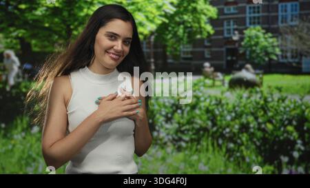 Femme portant un débardeur blanc tenant les mains sur la poitrine et souriant avec les yeux fermés dans le bâtiment ; satisfaction de gratitude. Banque D'Images