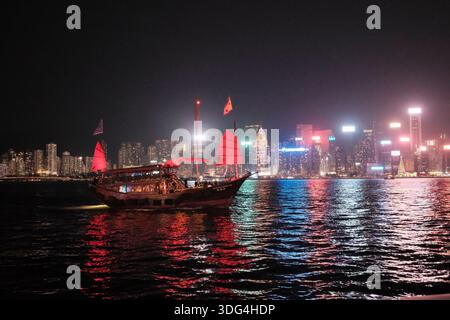 Une jonque chinoise traditionnelle traverse le port Victoria la nuit, avec les gratte-ciel illuminés de Hong Kong qui se reflètent dans l'eau derrière Banque D'Images