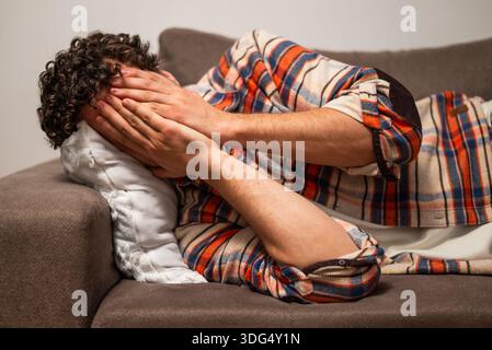 Jeune homme épuisé et déprimé tenant la tête dans les mains et allongé sur le canapé à sa maison. Banque D'Images