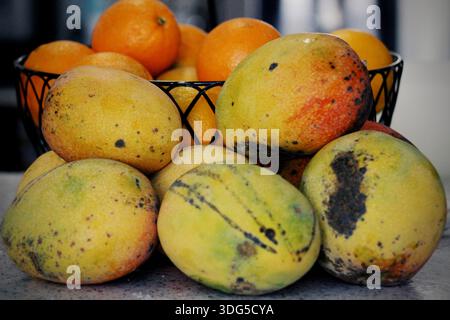 Pile de mangues mûres fraîches avec un bol d'oranges en arrière-plan, affichage coloré de fruits tropicaux. Banque D'Images