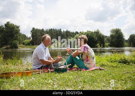 charmant couple de personnes âgées au lac ayant un pique-nique, faisant un toast sur la rive du lac un jour d'été. Staycations, voyage hyper-local, sortie en famille, escapade. || autorisation du modèle Banque D'Images