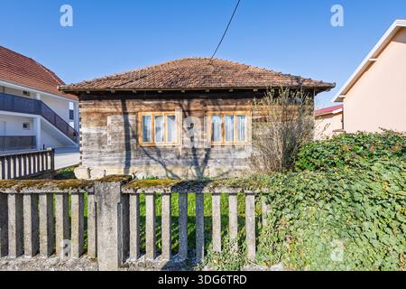 Un petit chalet en bois ancien se trouve à côté d'une clôture en béton mousseline, avec du lierre envahi par la végétation et des arbustes. Les maisons modernes donnent sur un quartier calme, sous un br Banque D'Images