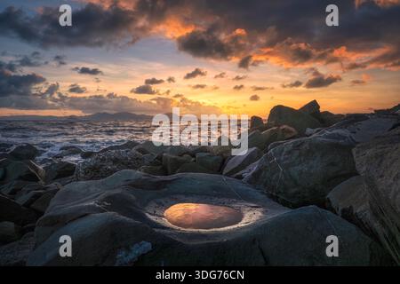 Coucher de soleil spectaculaire sur la côte rocheuse de Piombino, Toscane, Italie. Petite flaque d'eau reflétant des nuages orange avec la silhouette de l'île d'Elbe sur le Banque D'Images