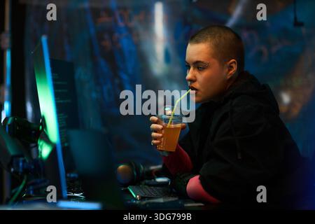 Jeune femme caucasienne adulte avec les cheveux courts assis au poste de travail, buvant du jus à travers la paille, tout en regardant un moniteur d'ordinateur dans une pièce sombre avec des éléments cyber visibles Banque D'Images