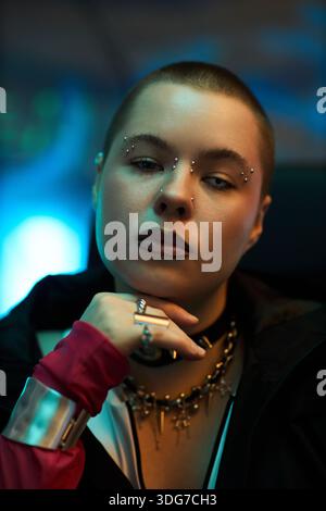Portrait d'une jeune femme caucasienne adulte avec la tête rasée et piercings faciaux assis à l'intérieur, menton reposant sur la main, portant des bijoux et regardant directement la caméra Banque D'Images