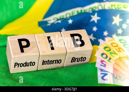 Le mot PIB, Produto Interno Bruto (produit intérieur brut) écrit sur des cubes en bois avec de l'argent réel brésilien en jaune, drapeau vert du Brésil. Brésil Banque D'Images