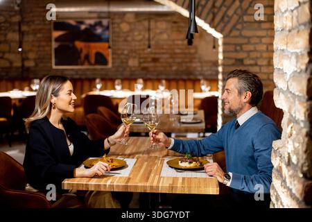 Un couple est assis à une table en bois dans un restaurant. Ils sourient et tiennent des verres de vin tout en partageant un repas ensemble. L'atmosphère est chaleureuse an Banque D'Images