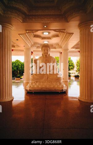 Une statue blanche de Bouddha se trouve en méditation à l'intérieur d'une salle de temple ornée, encadrée par des colonnes et des plafonds décoratifs, photographiée symétrie. Fév 2025 Banque D'Images