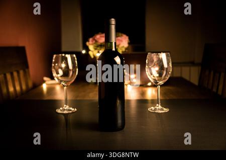 Bouteille de vin et deux verres sur une table avec des lumières bokeh chaudes et des fleurs en arrière-plan, scène de dîner intime. Banque D'Images