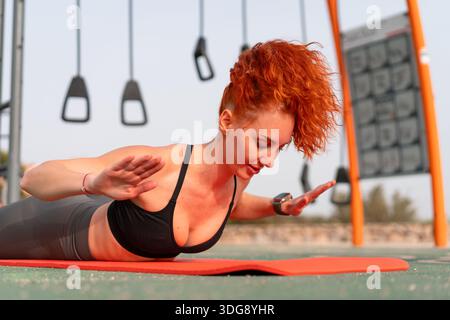 Fit femme rousse dans les vêtements de sport faisant de l'exercice au sol sur le tapis de yoga à la salle de gym extérieure pendant l'entraînement du matin, en maintenant un style de vie actif et sain avec concentration et force Banque D'Images