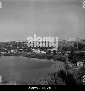 Slavyansk, région de Donetsk, Ukraine, URSS. Archive photo en noir et blanc des années 1980 montrant une vue panoramique du lac Liman 2 avec pour toile de fond un nouveau quartier résidentiel à plusieurs étages. L'image capture le contraste entre le paysage naturel tranquille du réservoir et l'architecture urbaine soviétique en développement. Au premier plan, la surface de l'eau calme reflète le ciel, avec une petite jetée métallique visible près du rivage envahi par la végétation. Des maisons privées et des arbres denses bordent le lac. Au loin, des rangées d'immeubles à panneaux bordent l'horizon, illustrant la croissance de l'indus paisible Banque D'Images