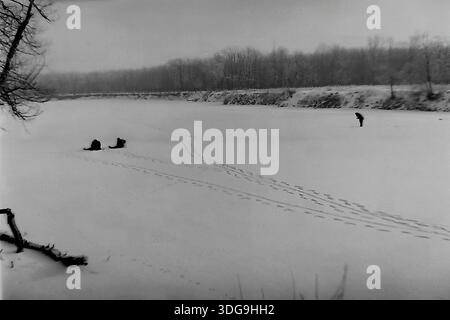 Slavyansk, région de Donetsk, Ukraine, URSS. Archive photo en noir et blanc des années 1960 capturant un vaste paysage hivernal sur une rivière gelée. La vaste surface blanche est parsemée de trois petites figures de pêcheurs de glace : deux sont assis près de leurs trous, tandis que l'une se tient plus loin. Des empreintes de pas distinctes sillonnent la neige. Au premier plan, un accroc sombre dépasse du banc de neige. Une ligne de forêt dense crée une toile de fond sous le ciel gris. Cette image atmosphérique illustre la solitude tranquille de la pêche hivernale dans la paisible région du Donbass. Banque D'Images